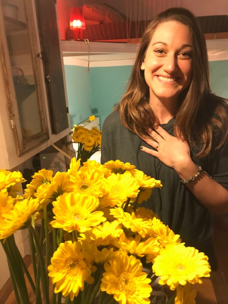 Yellow daisies are in the foreground and the background as I stand with my newly engaged hand on my chest featuring my ring.