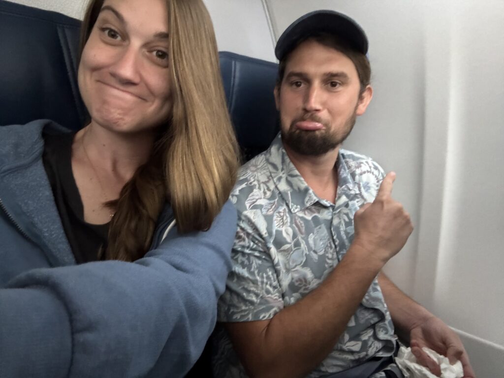 A couple sits in a row of an airplane with no window. They have silly pouty faces on and point to the lack of window.